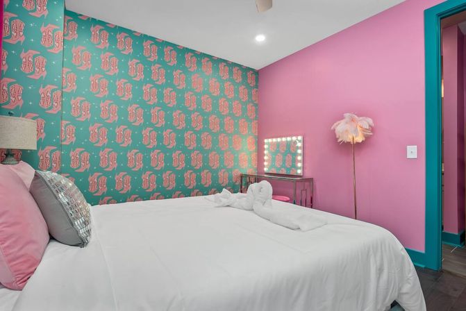 Vibrant modern bedroom interior with bright pink walls and teal patterned accent wall, white bed with pink and metallic pillows, mirrored vanity with bulb lights, feather floor lamp and teal door trim.
