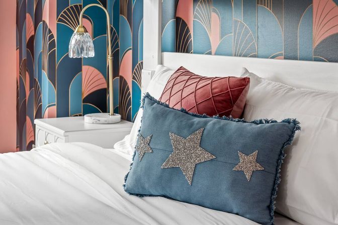 Close-up of a stylish bedroom: white bed with a blue star-appliqué throw pillow and pink quilted cushion, art-deco teal and blush wallpaper, brass bedside lamp on a white nightstand.