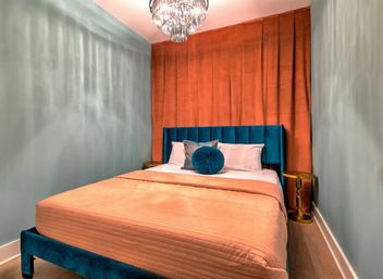 Stylish small bedroom with teal velvet headboard, coral bedspread and matching curtains, round teal accent pillow, gold side tables and crystal chandelier