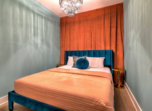 Stylish small bedroom with teal velvet headboard, coral bedspread and matching curtains, round teal accent pillow, gold side tables and crystal chandelier