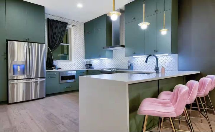 Modern kitchen with deep green cabinets, white quartz island, three pink velvet bar stools, stainless steel French-door refrigerator, white scalloped tile backsplash, black gooseneck faucet and gold pendant lights over light hardwood floors.