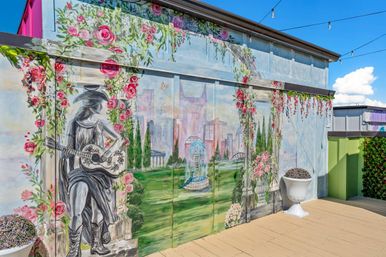 Urban rooftop mural of a seated guitarist in a cowboy hat framed by climbing pink roses and columns, revealing a painted city skyline, fountain and green park; wooden deck with string lights and white planters in the foreground.