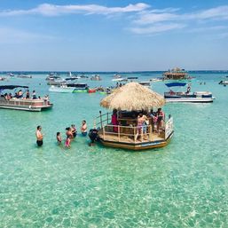 Floating tiki bar in crystal-clear turquoise water with people wading and pontoon boats nearby under a sunny blue sky — lively tropical beach party scene.