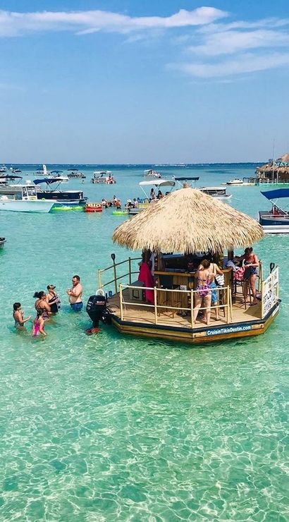 Floating tiki bar in crystal-clear turquoise water with people wading and pontoon boats nearby under a sunny blue sky — lively tropical beach party scene.