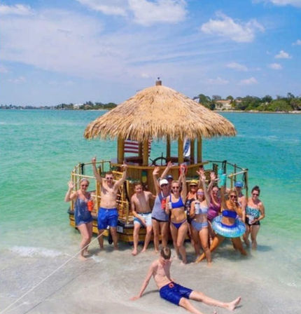 Group of people in swimsuits waving from a thatched-roof floating tiki raft beached on a sunny tropical shoreline with clear turquoise water.