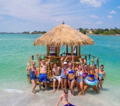 Group of people in swimsuits waving from a thatched-roof floating tiki raft beached on a sunny tropical shoreline with clear turquoise water.