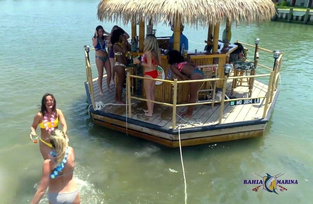 Group of women in swimsuits partying at a thatched-roof floating tiki bar anchored in calm, sunny marina waters, with some dancing on deck and others wading nearby.