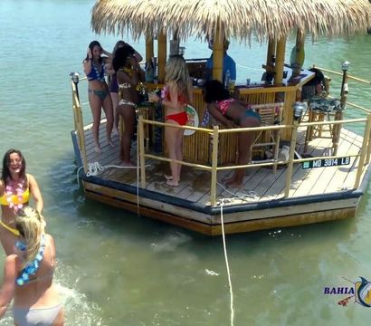 Group of women in swimsuits partying at a thatched-roof floating tiki bar anchored in calm, sunny marina waters, with some dancing on deck and others wading nearby.