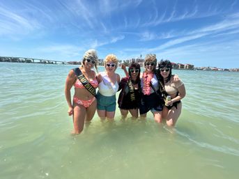 Five friends wearing novelty wigs, heart-shaped sunglasses, sashes and swimsuits standing waist-deep in shallow green bay water with a bridge and waterfront buildings under a bright blue sky