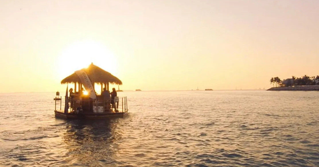 Thatched-roof floating tiki bar raft silhouetted against a golden sunset over calm tropical ocean, with a distant palm-lined coastline