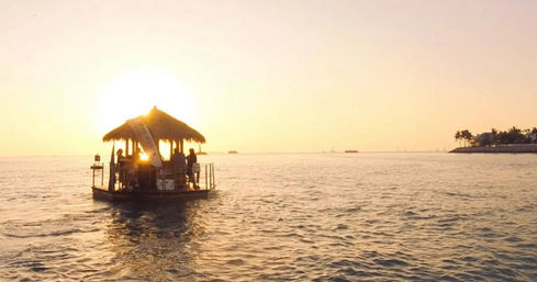 Thatched-roof floating tiki bar raft silhouetted against a golden sunset over calm tropical ocean, with a distant palm-lined coastline