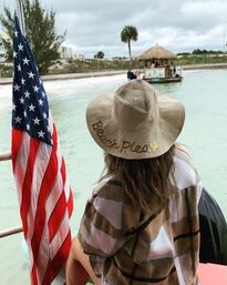Person wearing a wide straw sun hat embroidered 'Beach Please' and a plaid cover-up sits on a boat deck beside an American flag, looking across a turquoise lagoon toward a palm tree and tiki hut on a cloudy beach day.