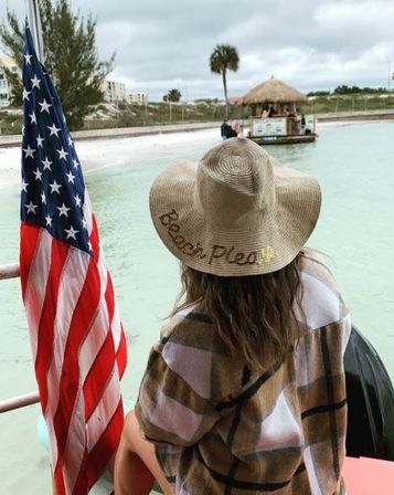 Person wearing a wide straw sun hat embroidered 'Beach Please' and a plaid cover-up sits on a boat deck beside an American flag, looking across a turquoise lagoon toward a palm tree and tiki hut on a cloudy beach day.