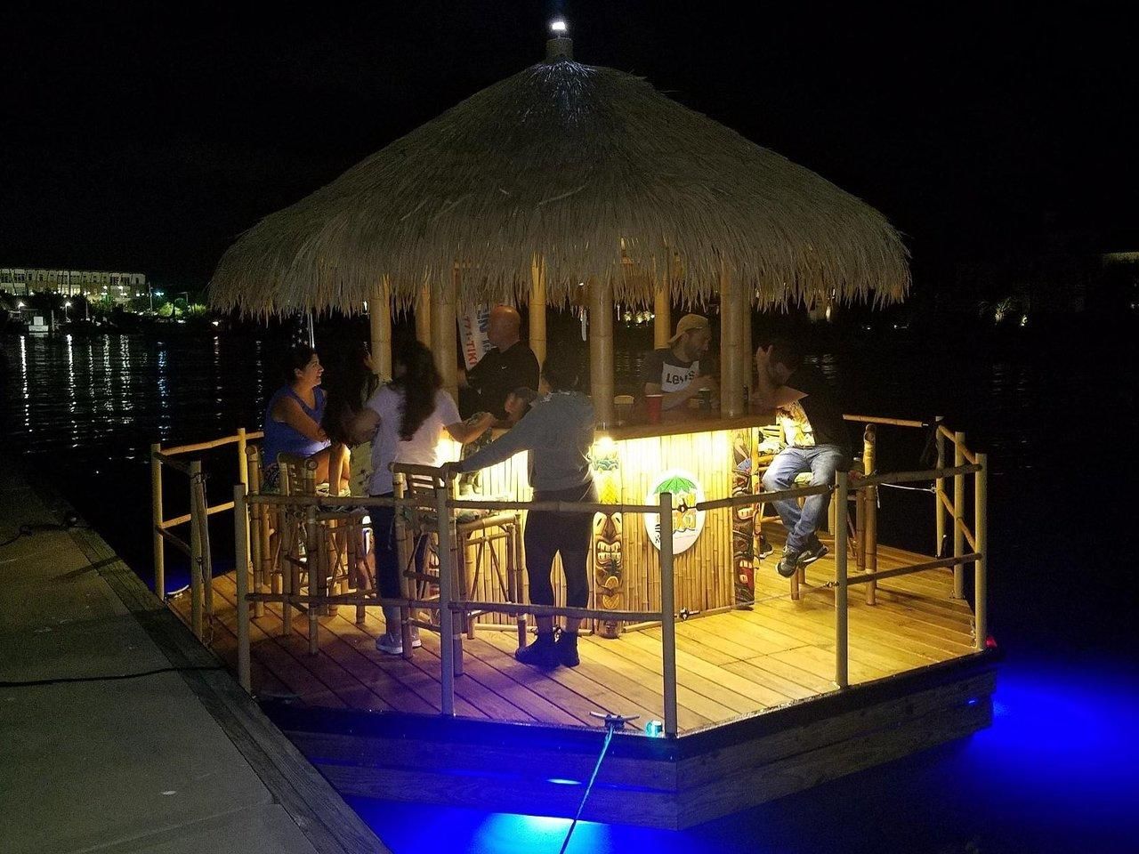 Nighttime floating tiki bar at a marina — wooden raft with thatched roof and bamboo railings, warm counter lighting over blue underwater LEDs, people socializing next to a dock.