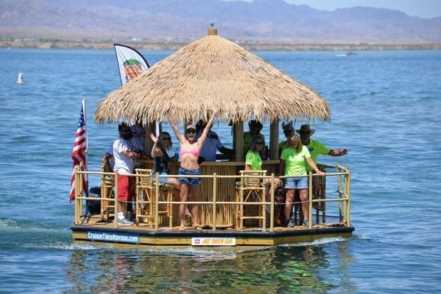 Tiki-style floating raft with a thatched roof carrying a cheerful group on a sunny blue lake with distant mountains and an American flag