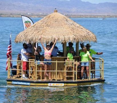 Tiki-style floating raft with a thatched roof carrying a cheerful group on a sunny blue lake with distant mountains and an American flag