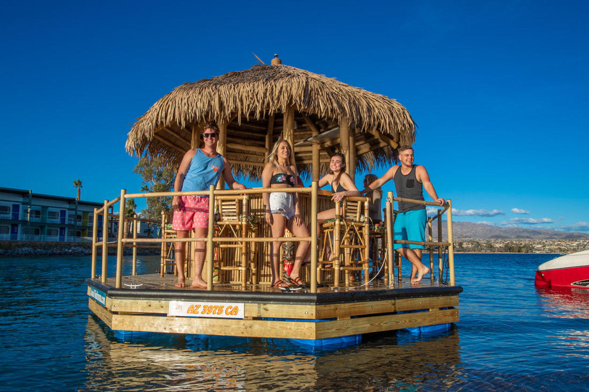 Four friends on a floating tiki-bar pontoon with a thatched roof, enjoying a sunny blue-sky day on a mountain-backed lake near a waterfront community.