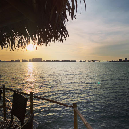 Golden sunset over a calm bay with a city skyline and arched bridge silhouetted in the distance, viewed from a waterfront tiki hut with woven chairs and bamboo railing.