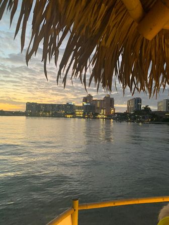 Tiki thatched roof silhouette framing a coastal city skyline at sunset, warm lights reflecting on calm harbor water.