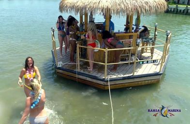 Women in swimsuits enjoying drinks on a thatched-roof floating tiki bar platform in calm marina waters on a sunny day.