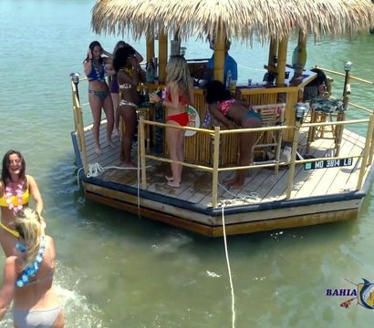 Women in swimsuits enjoying drinks on a thatched-roof floating tiki bar platform in calm marina waters on a sunny day.