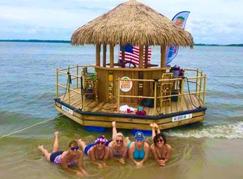 Group of five people in swimsuits lounging in shallow water in front of a thatched-roof floating tiki bar raft with an American flag off a calm shoreline