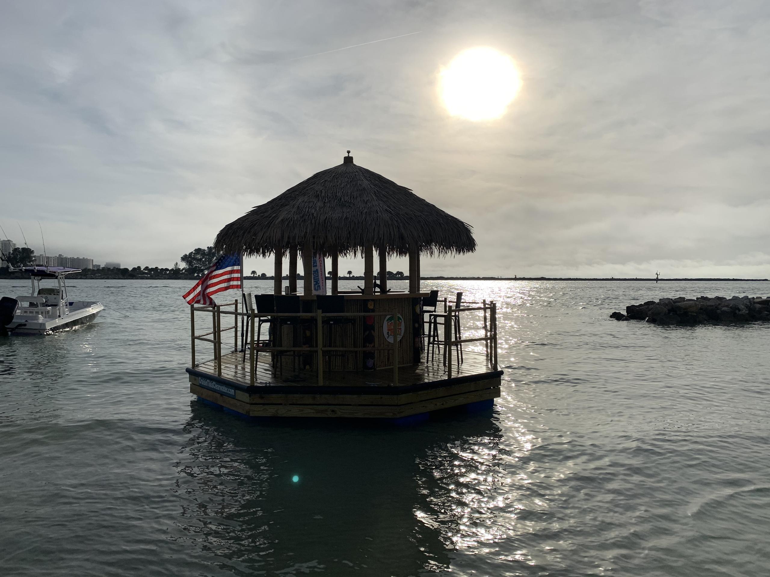 Thatched-roof floating tiki bar on a calm coastal bay, American flag and small boat nearby, bright low sun sparkling on the water.