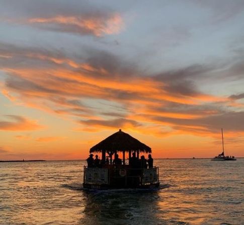 Silhouetted tiki-roofed boat of people cruising on calm ocean as vibrant orange-pink sunset paints the sky, with a distant catamaran on the horizon.