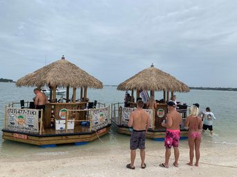 Two thatched-roof floating tiki bars moored just offshore at a sandy Florida Gulf Coast beach, people in swimsuits boarding and chatting, calm shallow water and cloudy sky.