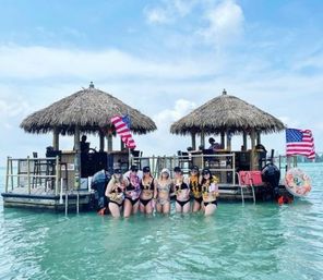 Group of women in swimsuits standing waist-deep in turquoise water in front of two thatched-roof floating tiki huts with American flags under a sunny tropical sky, enjoying a boat-bar outing.