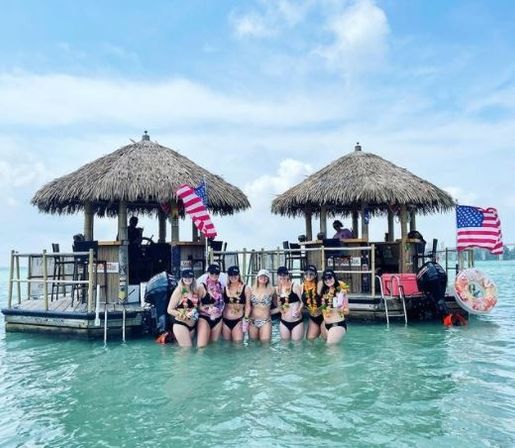 Group of women in swimsuits standing waist-deep in turquoise water in front of two thatched-roof floating tiki huts with American flags under a sunny tropical sky, enjoying a boat-bar outing.