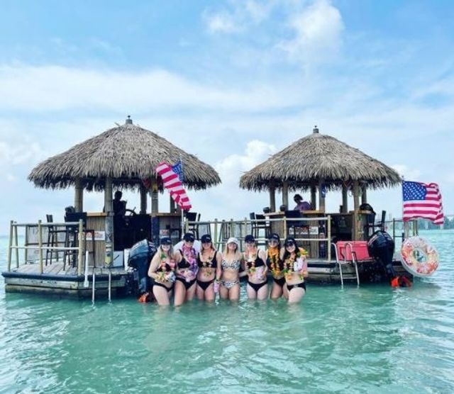 Group of women in swimsuits standing waist-deep in turquoise water in front of two thatched-roof floating tiki huts with American flags under a sunny tropical sky, enjoying a boat-bar outing.