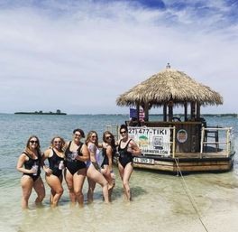Seven women in swimsuits posing waist-deep in clear turquoise water beside a thatched-roof floating tiki bar at a sunny tropical beach with a small island on the horizon.