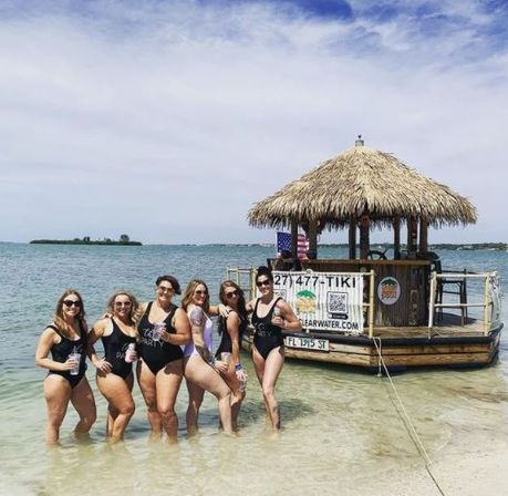 Seven women in swimsuits posing waist-deep in clear turquoise water beside a thatched-roof floating tiki bar at a sunny tropical beach with a small island on the horizon.