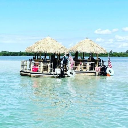 Two thatched-roof floating tiki bars with stools and American flags bobbing on turquoise coastal water under a sunny blue sky