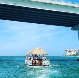 Group on a floating tiki bar boat with a thatched roof cruising turquoise coastal waters under a bridge on a sunny day