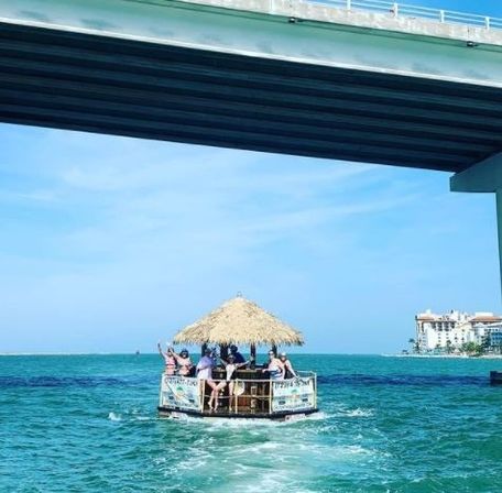 Group on a floating tiki bar boat with a thatched roof cruising turquoise coastal waters under a bridge on a sunny day