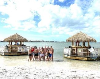 Group of friends posing in shallow turquoise water between two floating tiki bars on a sunny tropical beach with a distant shoreline and partly cloudy sky