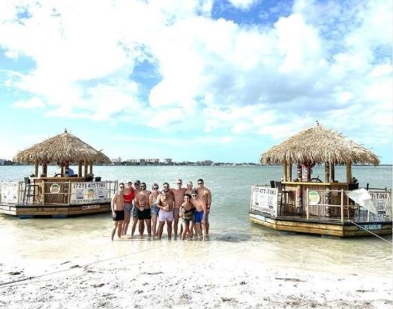 Group of friends posing in shallow turquoise water between two floating tiki bars on a sunny tropical beach with a distant shoreline and partly cloudy sky