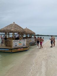 Two thatched-roof floating tiki bars anchored at a sandy beach sandbar, people wading in shallow green water under an overcast sky.