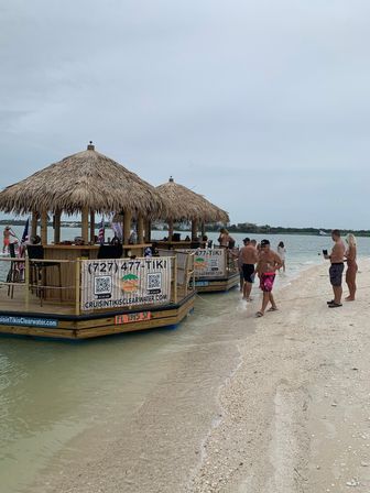 Two thatched-roof floating tiki bars anchored at a sandy beach sandbar, people wading in shallow green water under an overcast sky.