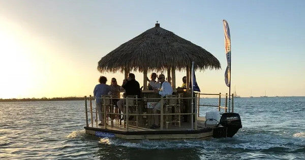 Group of people enjoying drinks under a thatched-roof floating tiki bar on a motorized pontoon at sunset over calm coastal waters.