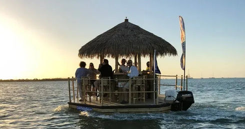 Group of people enjoying drinks under a thatched-roof floating tiki bar on a motorized pontoon at sunset over calm coastal waters.