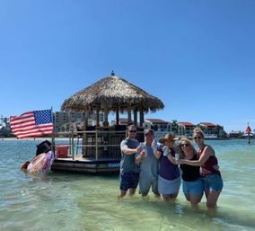 Five friends wading in clear shallow water, cheering with canned drinks beside a floating tiki bar with a thatched roof and an American flag, low-rise waterfront buildings and bright blue sky behind them.