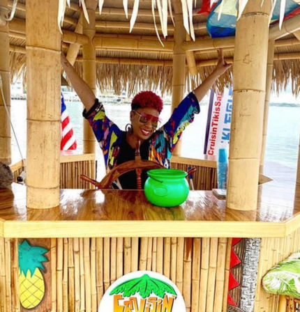 Cheerful person with red hair and colorful shirt raising arms behind a bamboo tiki hut bar on the water, posing at a wooden ship wheel with a bright green bowl on the counter, pineapple decor and an American flag in the background.