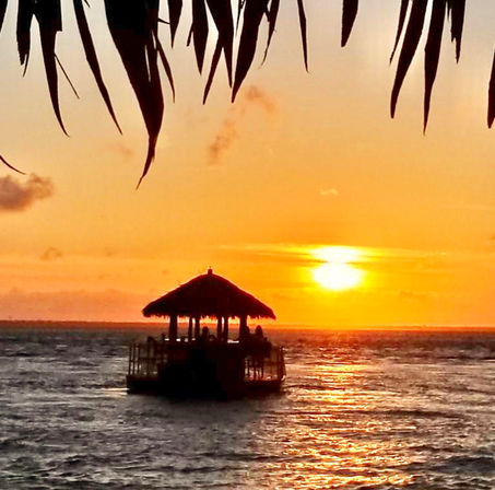 Golden tropical sunset over the ocean with a silhouetted thatched-roof tiki hut on a floating platform, framed by palm fronds