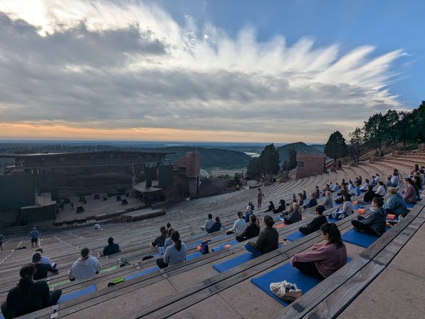 Sunset group yoga on mats at a large outdoor stone amphitheater on a rocky hillside, attendees seated on tiered benches facing a stage with sweeping cloud-filled sky and distant valley view.