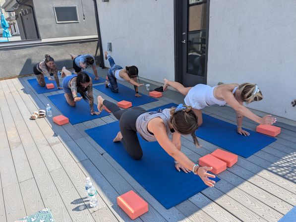 Small group rooftop yoga class on a sunny urban deck, women balancing in bird-dog pose on blue mats with coral yoga blocks and water bottles nearby.