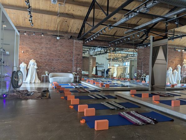 Industrial brick loft studio with exposed wooden beams set up for yoga: rows of blue mats, orange blocks, patterned blankets on polished concrete, large wall mirrors, bridal mannequins in white gowns and a cozy sofa.