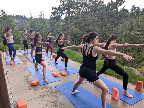 Outdoor group yoga class doing Warrior II on blue mats with orange blocks on a patio overlooking pine-covered mountains